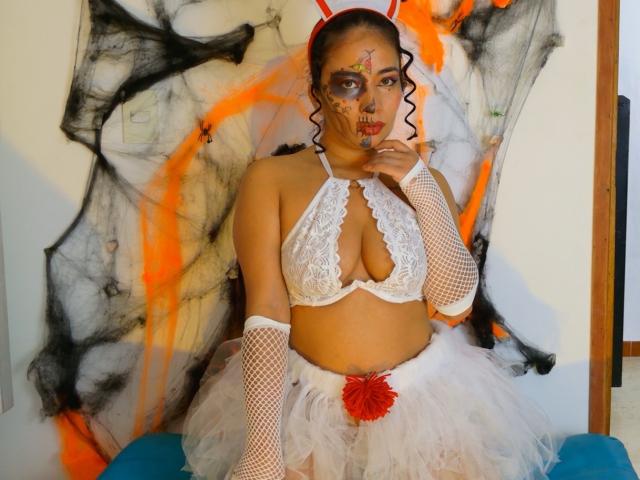 AmaraMartellI stands in the center, wearing a white lace halter top with a deep cleavage, white fishnet arm sleeves, and a white tutu skirt with a red flower. She has dark curly hair, red bunny ears, and face paint with orange and black patterns. She touches her chin with one hand. The background features a black and orange abstract painting. Her skin tone is medium brown. The image has a playful, provocative feel.