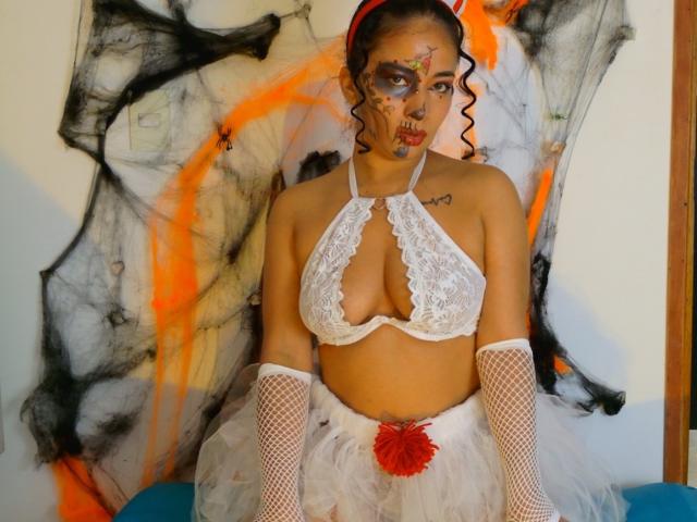 AmaraMartellI is centered in the image, wearing a white lace halter top with a deep cleavage, exposing her medium-sized breasts. She has white fishnet gloves and a white tutu with a red floral accent. Her face is painted with abstract, colorful designs, and she has curly dark hair with a red headband. A small tattoo is visible on her left shoulder. The background has black, orange, and white abstract patterns. Her expression is neutral.