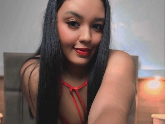 MissOrtizz with long black hair, red lipstick, and a red strappy top, leaning forward, showing her bare shoulders and a slight smile. Her boobs are partially visible.