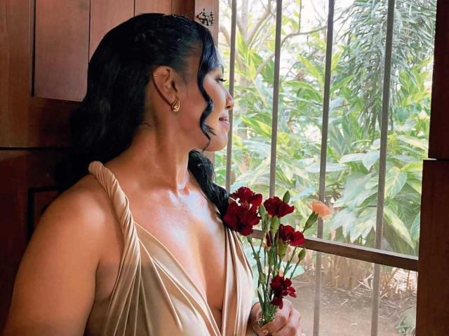 MissOrtizz with tan, deep-cut dress, holding red flowers, standing by window, black wavy hair, earrings, looking outside, wooden door, green plants outside, soft sunlight on her skin.