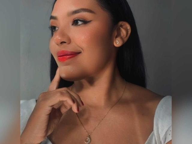 MissOrtizz with dark hair, red lipstick, and earrings, touching her chin, wearing a white off-shoulder top, and a gold necklace, smiling softly. Her tits are partially visible.