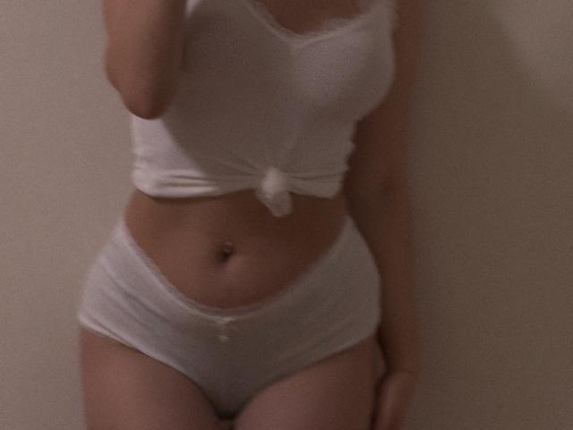 SugarrX wearing a white, tied-up crop top and white, low-rise panties, showing her bare midriff and a small belly button, with a soft, blurry, dimly-lit photo.