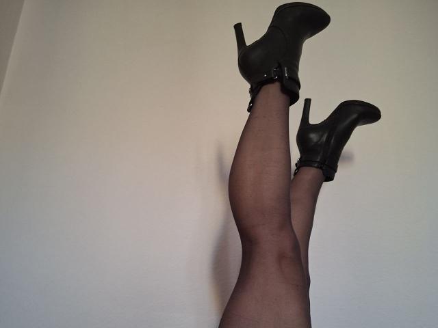 A close-up of SexiOxi's legs, wearing black high-heeled boots with straps, standing against a plain white wall. The legs are covered in sheer black stockings, showing subtle patterns. The boots have a glossy texture and the heels are pointed. SexiOxi's legs are slightly apart and angled upwards, creating a dynamic composition. The image focuses on the contrast between the glossy boots and the sheer stockings.