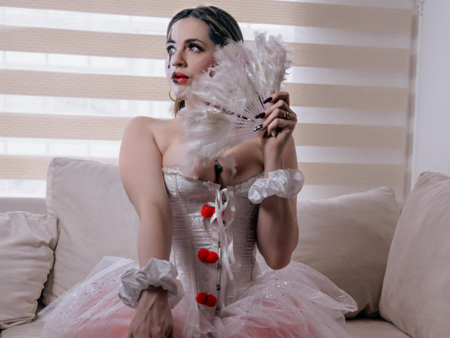 PaulettWatsoon with white feather fan, wearing a strapless pink corset with red buttons, white ruffled wrists, and pink tulle, sitting on a beige couch, looking up.