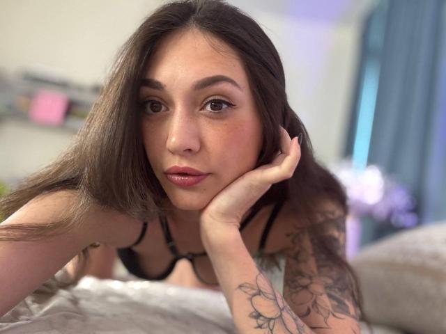 MoniFoxy with long brown hair, brown eyes, and pink lips, wearing black lingerie, head resting on hand, tattoo on arm, looking at camera, lying on bed.