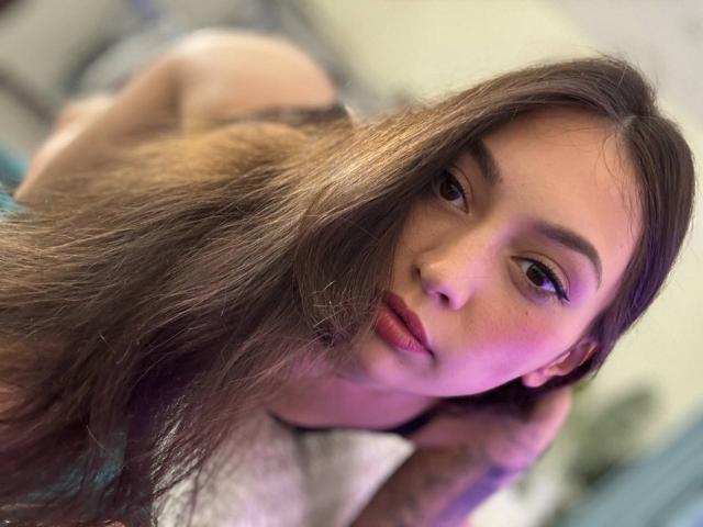 MoniFoxy's face with long brown hair, pink-lit skin, and subtle makeup, looking at the camera with a slight pout, her small tits and bare ass in the background.