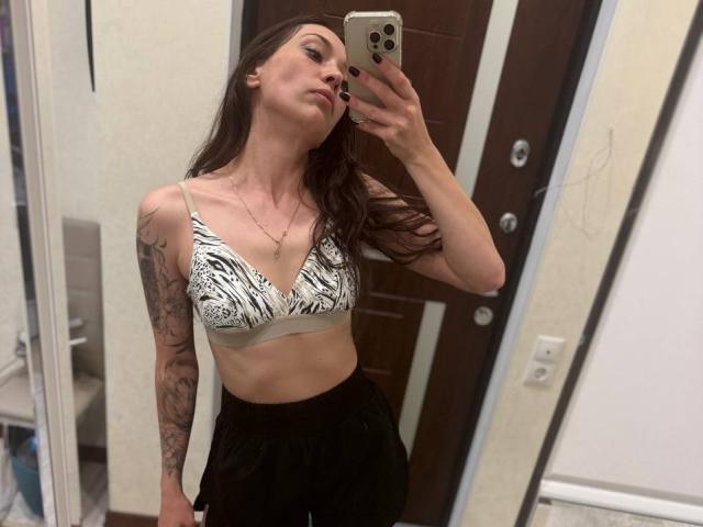 MoniFoxy with long brown hair, wearing a zebra-patterned bra and black pants, holding a phone, showing tattoos on her right arm, standing in front of a mirror.