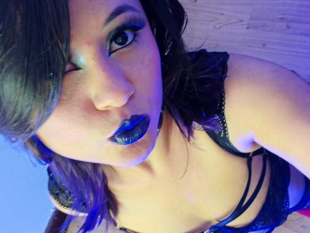 CassieJones with dark blue lipstick, winking, wearing black lace lingerie, showing her small tits and cleavage, on wooden floor, in a purple-blue lit room, looking at camera.