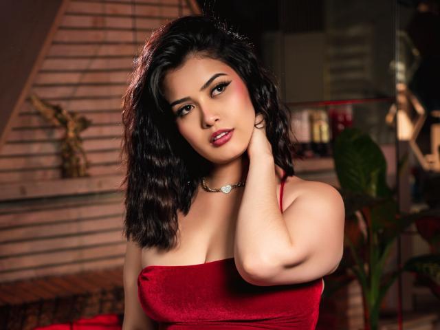 MiaWoodss with wavy black hair, red velvet dress, and a necklace, touching her neck, looking at the camera with red lipstick, soft makeup, and a sexy, confident expression.