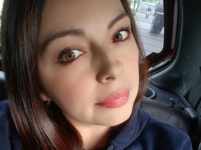 Seher's face with wide brown eyes, shiny pink lips, and dark brown hair, wearing a blue hoodie, sitting in a car, looking slightly to the side.