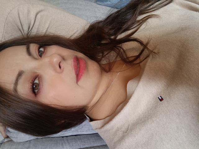Seher with brown hair, green eyes, and pink lipstick, wearing a beige sweater with a small red and blue logo, lying down, smiling softly.