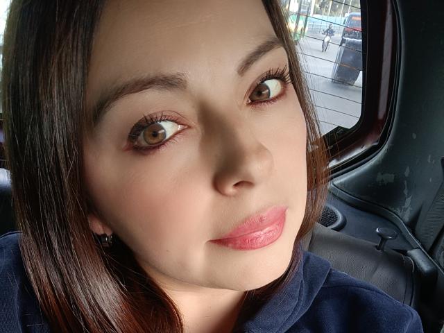 Seher has brown eyes, glossy pink lips, and straight dark brown hair. She's wearing a black hoodie, sitting in a car with a window behind her.