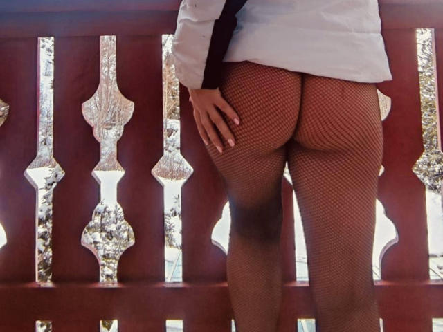 ReineDuVice's butt in red fishnet stockings, wearing a white shirt, standing with her back to the camera, hand on her right ass cheek, on a red balcony.