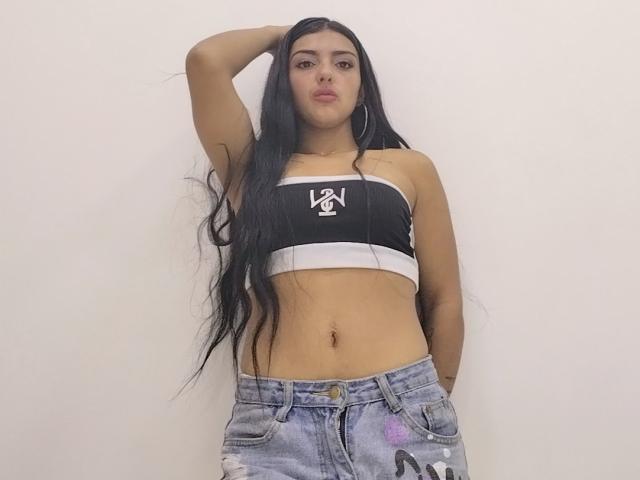 Naomhy with long black hair, wearing a black and white strapless top, showing her flat stomach, and blue distressed jeans, hand in pocket, arm raised, pouty expression.