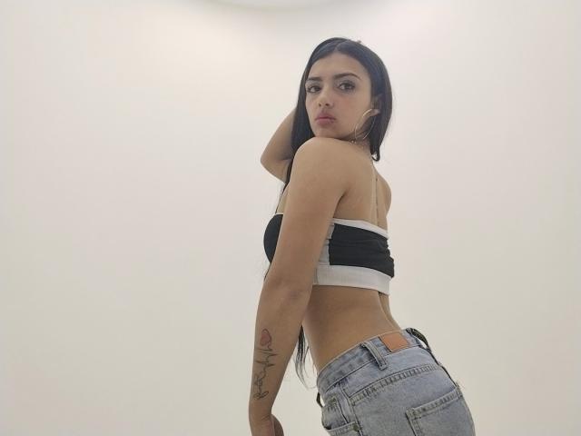 Naomhy with long black hair, small black and white tube top, showing her back and ass, wearing light blue jeans, has a tattoo on her right arm, looking over shoulder.