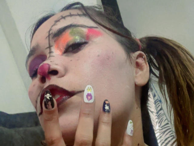 SophiaQueens mit auffälligem Make-up, einer rosafarbenen Nase, roten Lippen und Tattoos, trägt Nageldesign in Schwarz und Weiß mit Charaktermotiven, hat braunes Haar zu einem Pferdeschwanz gebunden und trägt einen Nasenring..