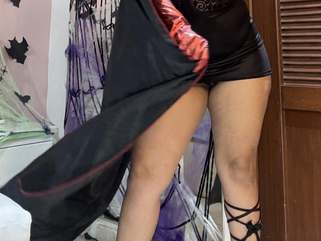 Close-up of SophiaQueens' lower body in a room. She wears a black, sheer mini skirt and black, shiny shorts, with black strappy heels. Her right leg is slightly raised, revealing her bare thighs. A black cape with red lining is partially visible, held by her red-gloved hand. The background includes a wooden door and a purple, star-patterned curtain. The image is slightly blurred. Her skin is light brown. The composition emphasizes her legs and the clothing.