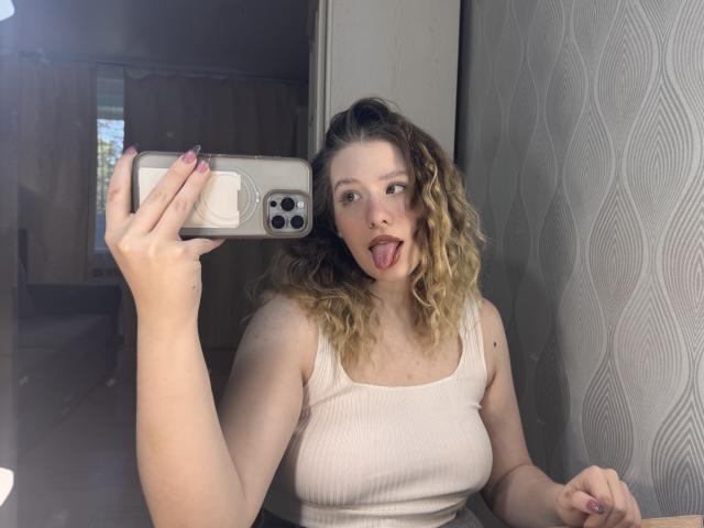 Tomioka69 con i capelli biondi ricci, la lingua fuori, che tiene un telefono, indossa un top bianco a coste, mostrando il seno, davanti a un muro con un motivo ondulato..