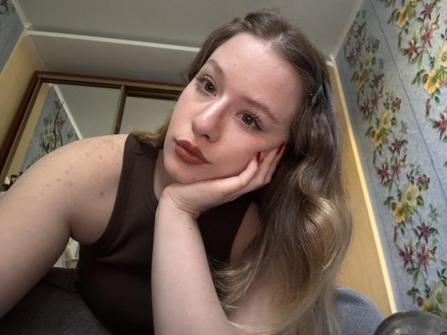 Tomioka69 con capelli castano ondulati, top marrone scuro senza maniche e rossetto rosso, con il mento appoggiato sulla mano, che guarda la fotocamera, mostrando lentiggini e un'espressione facciale delicata..