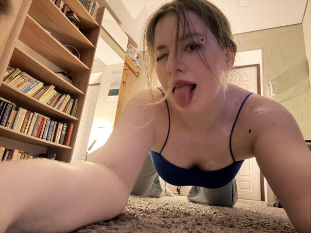 Tomioka69, tongue out, blue bra, on carpet, knees up, bookshelf left, wooden door, green wall, blonde hair, nose piercing, fair skin, selfie, looking at camera, messy hair.