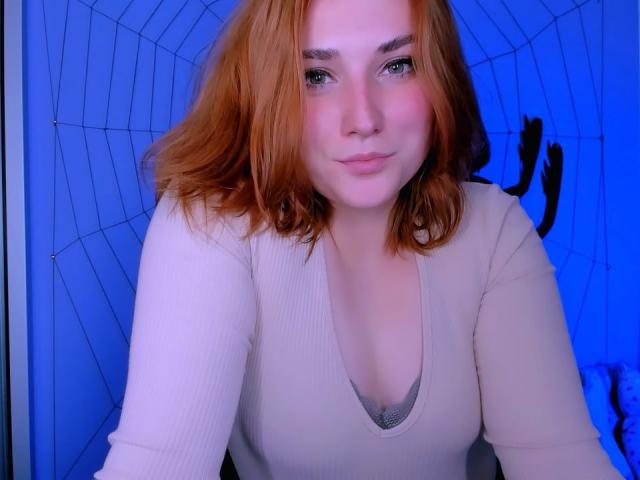 CaseyMeli with red hair, pale skin, and green eyes, wearing a beige ribbed top, has small tits and a cute, pouty face, looking at the camera.