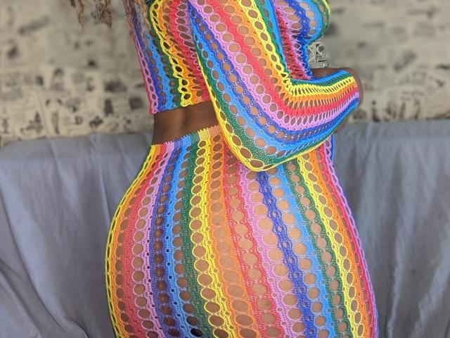 A close-up of OpheliaVayne's side, wearing a vibrant, rainbow-colored crochet dress. The dress features multiple colorful patterns, including yellow, pink, blue, and green, with circular lace patterns. A brown belt cinches the waist. OpheliaVayne's curvy, hourglass figure is emphasized by the dress's form-fitting design. The dress's texture is detailed and intricate, contrasting with the smooth, grey fabric in the background. The focus is on the dress's colorful, patterned design and OpheliaVayne's shape.