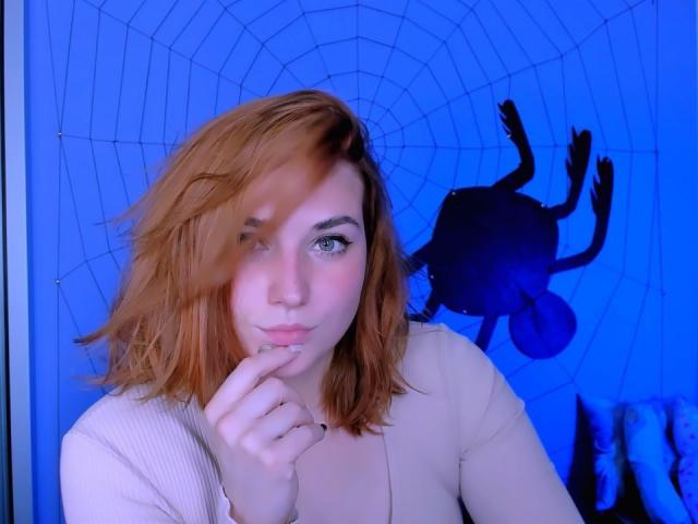 CaseyMeli with pinkish-red hair, blue eyes, and a cute face, touching her lips, wearing a beige top, in front of a blue spider web and shadow.