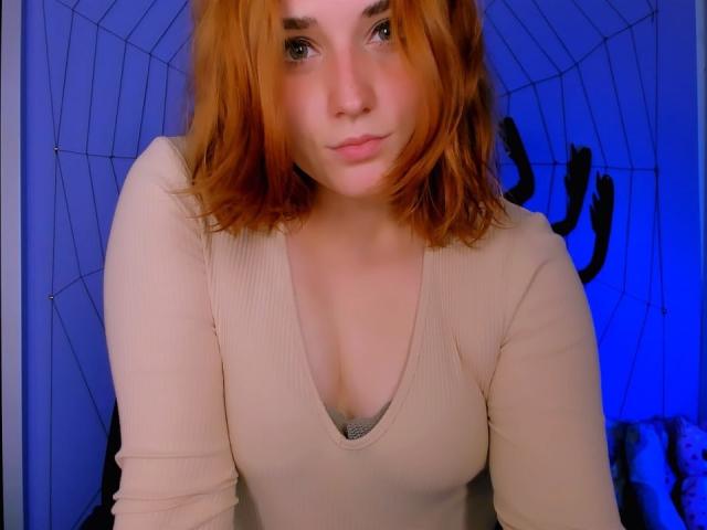 CaseyMeli with red hair, wearing a beige ribbed top, showing small tits in a deep V-neck, looking at the camera with a neutral expression. Blue lighting in background.