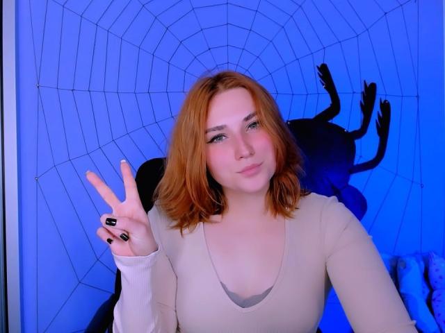 CaseyMeli with red hair, pale skin, black nail polish, wearing a beige top, making a peace sign, blue-lit background, spiderweb silhouette, subtle smile.