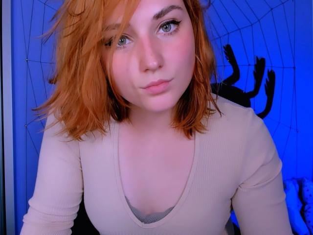 CaseyMeli with red hair, blue eyes, and a pout, wearing a beige top, has small tits and a cute face, looking at the camera in a blue-lit room.