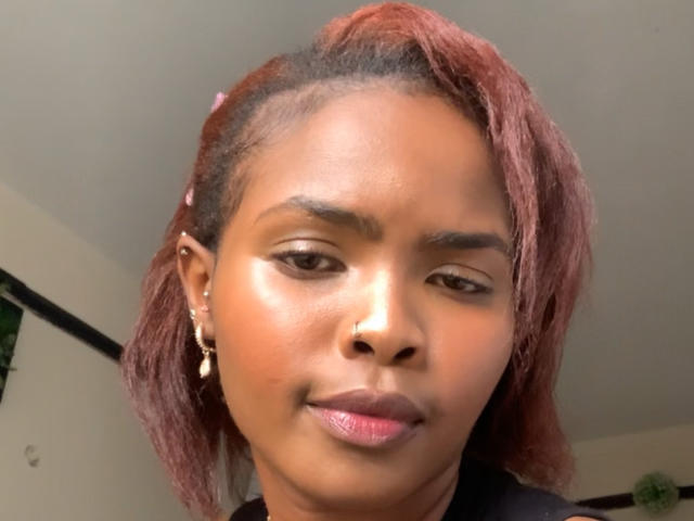 Niiia has dark brown skin, reddish-brown hair with pink tips, a nose ring, and gold earrings, smiling with pink lipstick, showing her shiny, smooth face.