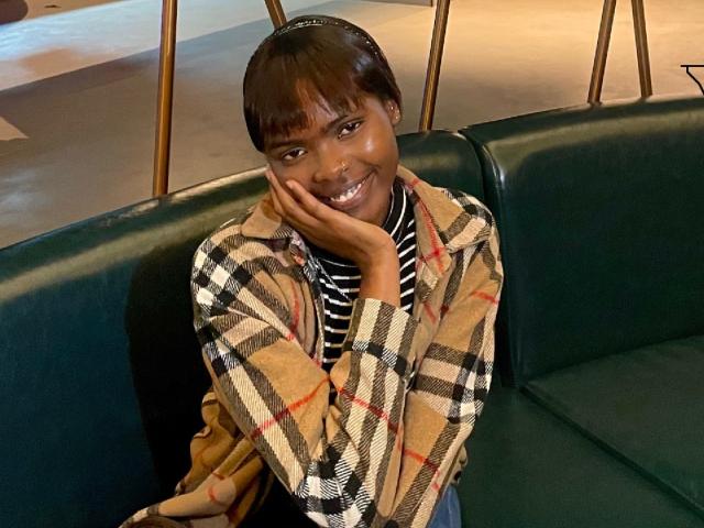 Niiia sits on a dark green leather bench, smiling with her right hand touching her cheek. She has a short, straight black hairstyle with bangs. She wears a tan and black plaid jacket over a black and white striped shirt. Her skin is dark, and she has a small nose ring in her right nostril. The lighting is warm, highlighting her cheerful expression. The bench has a modern, minimalist design with wooden legs visible in the background.