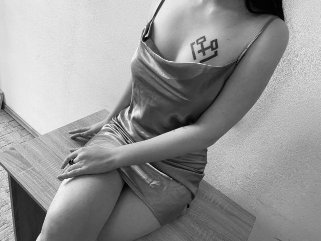 Black-and-white photo of MarriAnni with small tits, wearing a shiny satin dress, sitting on a wooden bench, showing a geometric tattoo above her right boob, legs crossed.