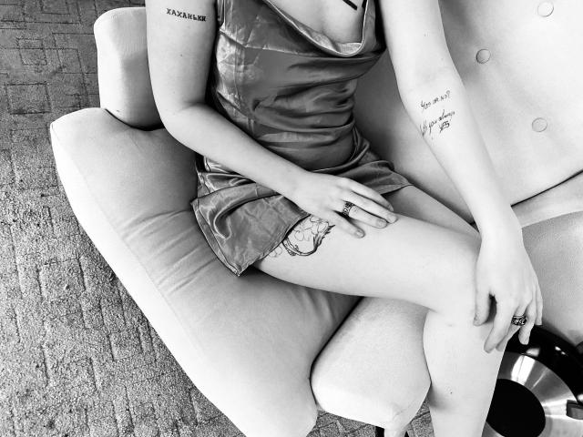 MarriAnni in a shiny, short, metallic dress, sitting on a chair, showing tattoos on her arm and thigh, crossed legs, ring on her hand, black and white image.