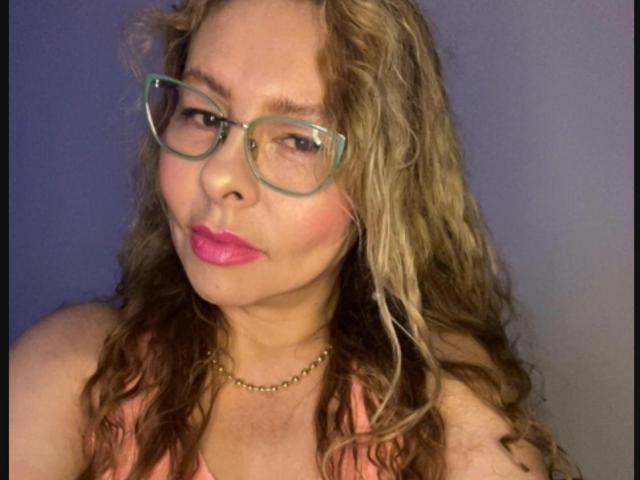 MiaAmoretti with wavy blonde hair, green glasses, pink lipstick, and a gold necklace, showing a slight pout. Boobs visible in a pink top. Medium skin tone.