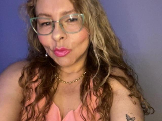 MiaAmoretti, con i capelli castano ricci, gli occhiali verdi, il rossetto rosa, la collana di perle, la maglietta rosa e un piccolo tatuaggio sulla spalla destra, che fa una smorfia..