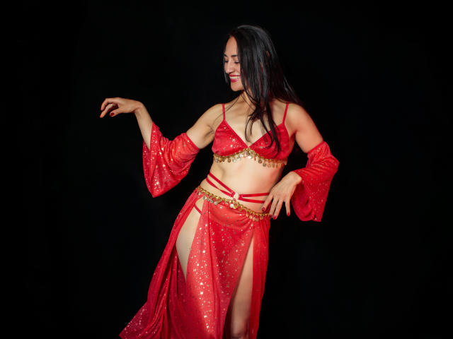 MiaMott with long black hair, red belly dancer outfit, gold trim, showing her tits in a red bra, and red sheer skirt with high slits, black background.