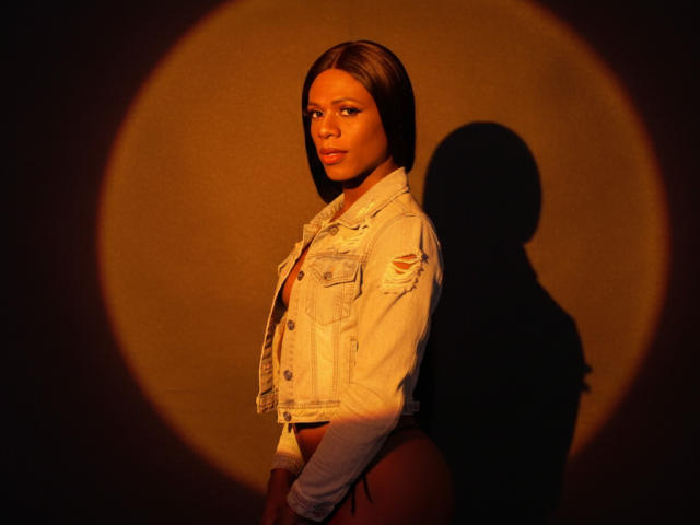 VenusMiller with short brown hair, wearing a white denim jacket, standing against a dark wall, shadowed, looking to the side, with a soft orange light on her face.