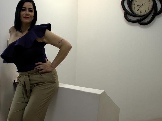 RebekaHunter stands in a corner, leaning on a white block. She has straight black hair, a confident smile, and wears a dark blue, one-shoulder top with ruffled edges and beige high-waisted pants. Her right hand rests on her hip. A tattoo is visible on her left upper arm. A round clock with a black frame is on the right wall. The image is sharp with neutral lighting. She has a light skin tone and is looking directly at the camera. Her makeup is subtle. The clock is at the top right.