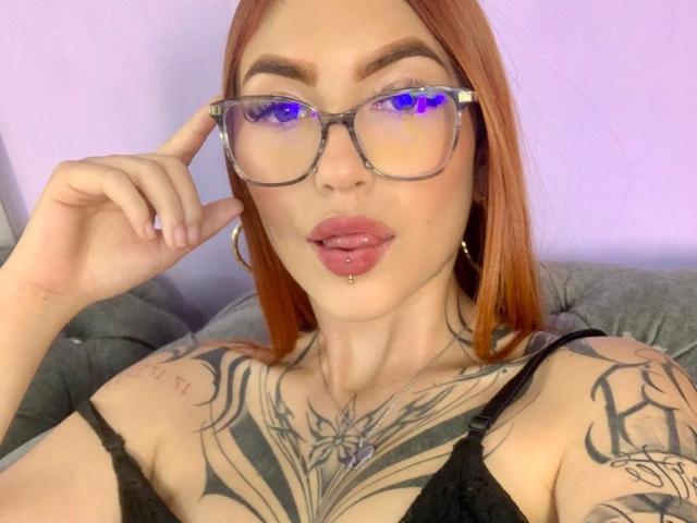 AntonellaBrith, avec des cheveux roux, des lunettes à monture bleue, une langue percée, un haut en dentelle noire, des tatouages sur la poitrine et les épaules, touchant ses lunettes, des lèvres roses, des boucles d'oreilles cerceaux, regardant l'appareil photo..