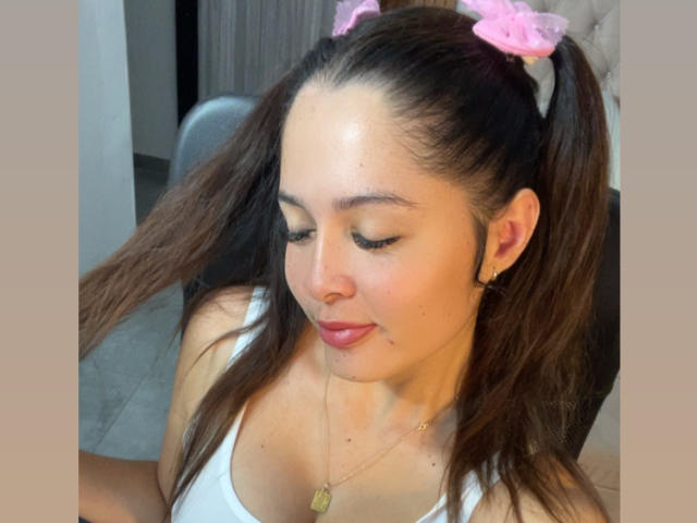 Gabrielafoxxy has brown hair in pigtails with pink scrunchies, wearing a white tank top, gold necklace, and earrings, looking down with closed eyes and a slight smile.