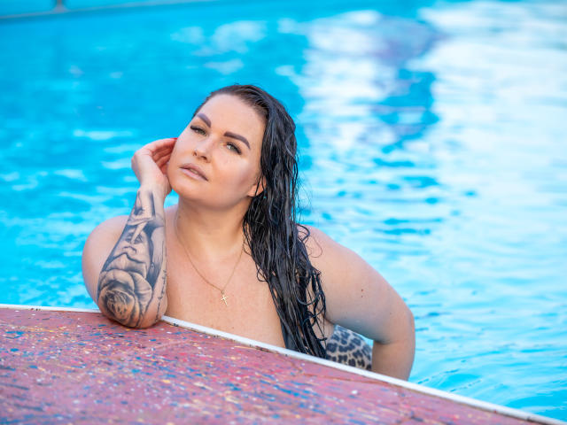 GeorgiaLerox with wet black hair, tattoos on her right arm, and a cross necklace, leaning on a pool edge, looking off to the side with a slight smile.
