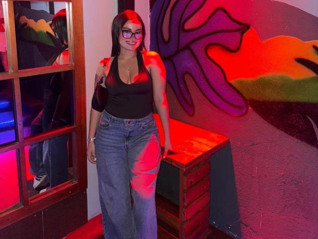 Miia Palmer, con un seno prosperoso, indossa una canotta nera, jeans blu e occhiali, ed è in piedi in una stanza con graffiti rossi e viola, tenendo in mano una borsa nera..