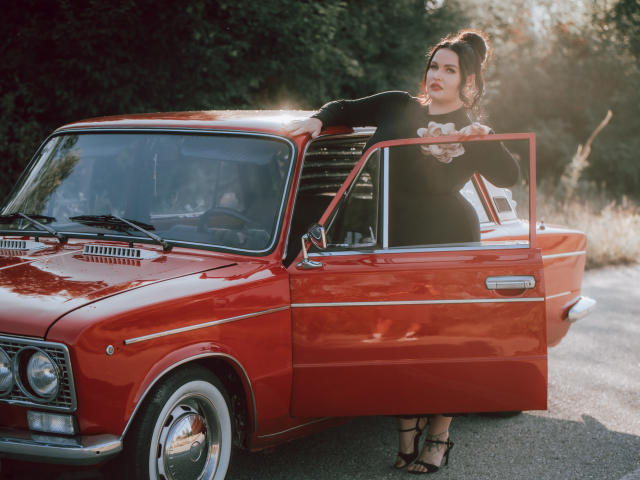 GeorgiaLerox with red lipstick, black top, and high heels leans out of a shiny red vintage car, holding a book, sunlight on her face, and her tits slightly visible.