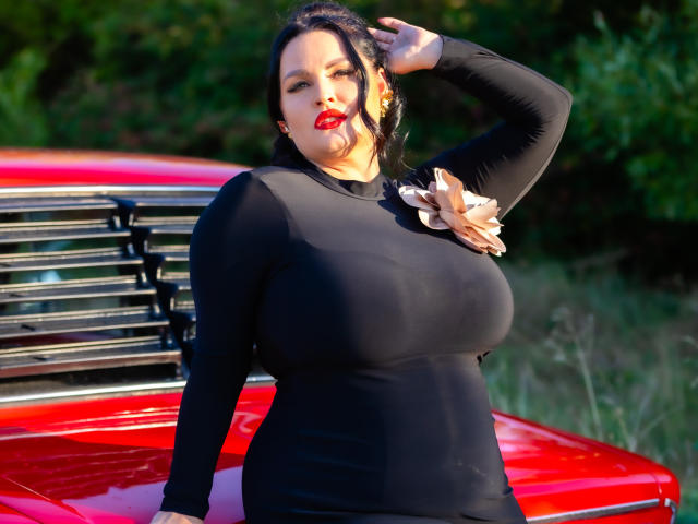 GeorgiaLerox with large tits, black long-sleeve dress, red lipstick, black hair, standing by a shiny red car, hand in hair, floral accessory on dress, green background.