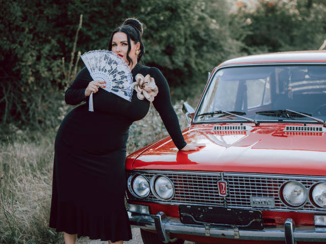 GeorgiaLerox with big tits, black dress, and hand fan, leaning on a red vintage car, holding a rope, dark hair in a bun, standing in grassy area.