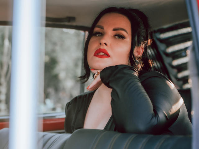 GeorgiaLerox with black hair, red lipstick, and black leather gloves, resting chin on hand, wearing a deep V-neck black top, sitting in a car, looking seductive.