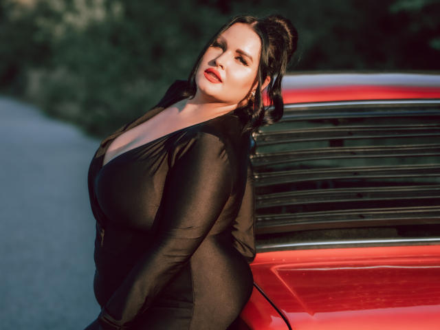 GeorgiaLerox with large tits, wearing a black, long-sleeve, deep V-neck, sitting against a red car, looking seductively to the side, dark wavy hair, red lipstick, sunlight.