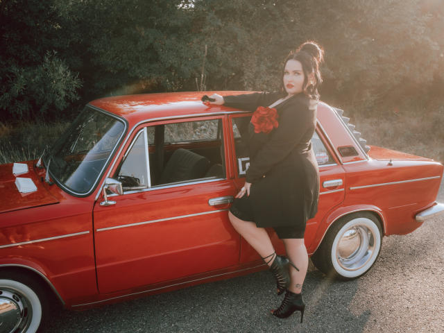 GeorgiaLerox leans against a shiny red classic car, wearing a black dress with a red rose, high heels, and black lace-up boots, with her ass and tits slightly visible.
