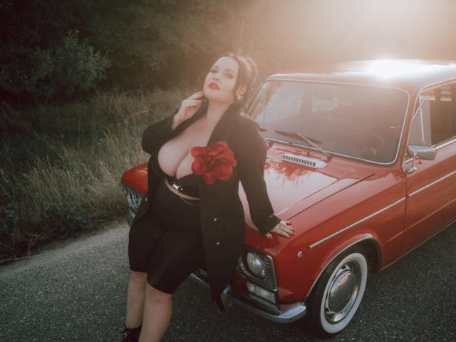 GeorgiaLerox with huge tits, wearing a black dress and jacket, red rose, leaning on a red classic car, sunlight, dark hair, red lipstick, standing on a gravel road.